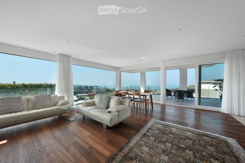 Modern living room with large windows offering a scenic view, featuring a cream-colored sectional sofa, a glass coffee table, a dining area with a wooden table and chairs, and a patterned rug on a wooden floor.