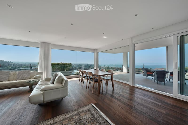 Modern living and dining area with large windows offering a scenic view, featuring a white sectional sofa, a wooden dining table with chairs, and a patterned rug on a wooden floor.