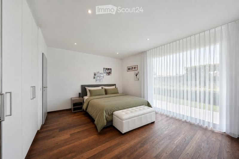 Modern bedroom with wooden flooring, large window with sheer curtains, bed with green bedding, nightstand, and a white leather stool.