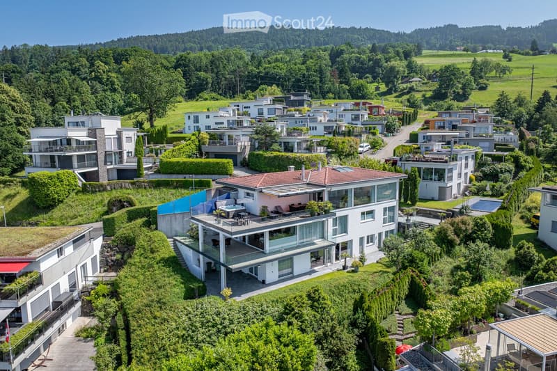 Aerial view of a residential area with multiple modern houses, surrounded by lush greenery and hills in the background.