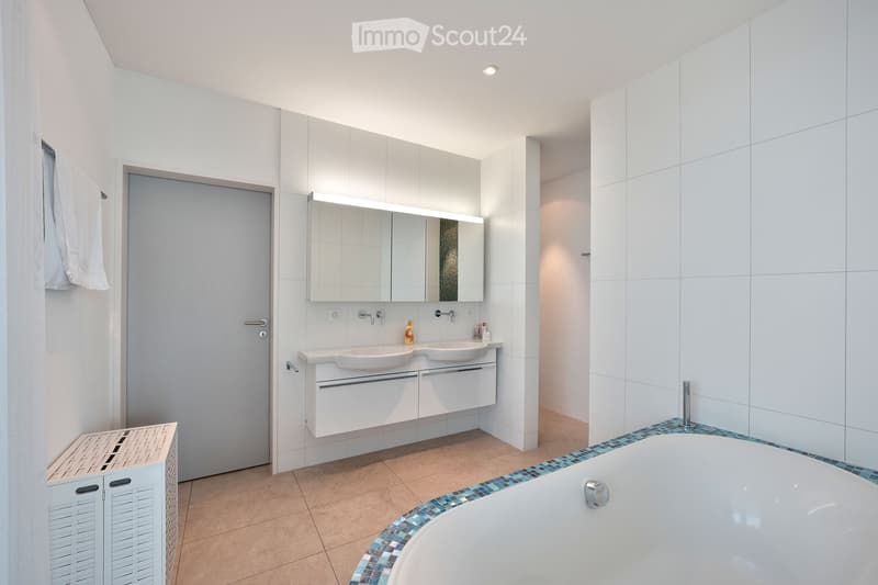 Modern bathroom with a white tiled wall, a bathtub with a mosaic tile border, a double sink vanity with a large mirror, and a towel hanging on the wall.