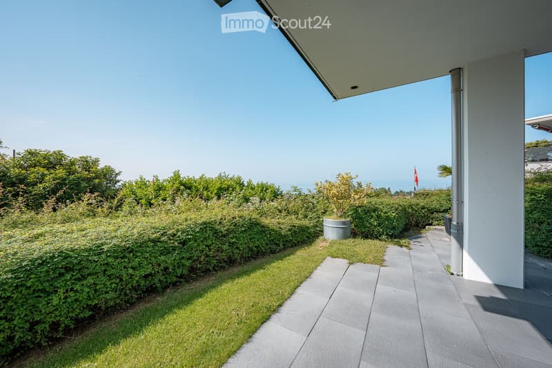 A well-maintained garden area with neatly trimmed hedges, a small grassy patch, and a paved pathway leading to a covered outdoor space. The background features a clear blue sky and distant water views.