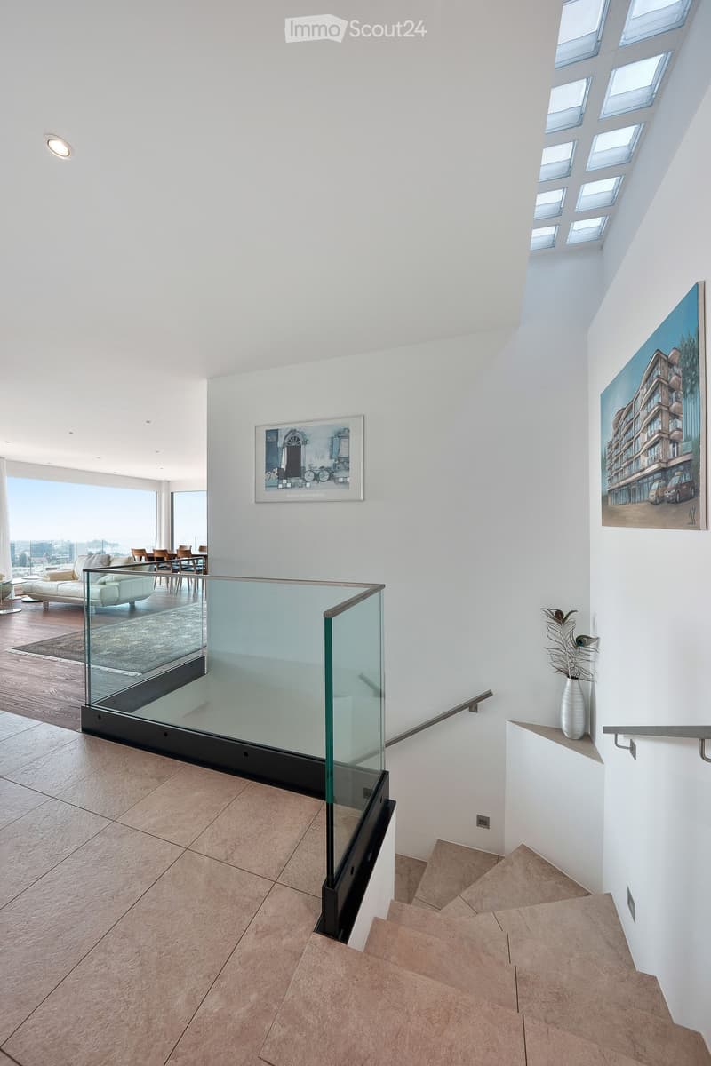 Modern hallway with a glass railing staircase, white walls, and a view of a living area with a sofa, dining table, and chairs.