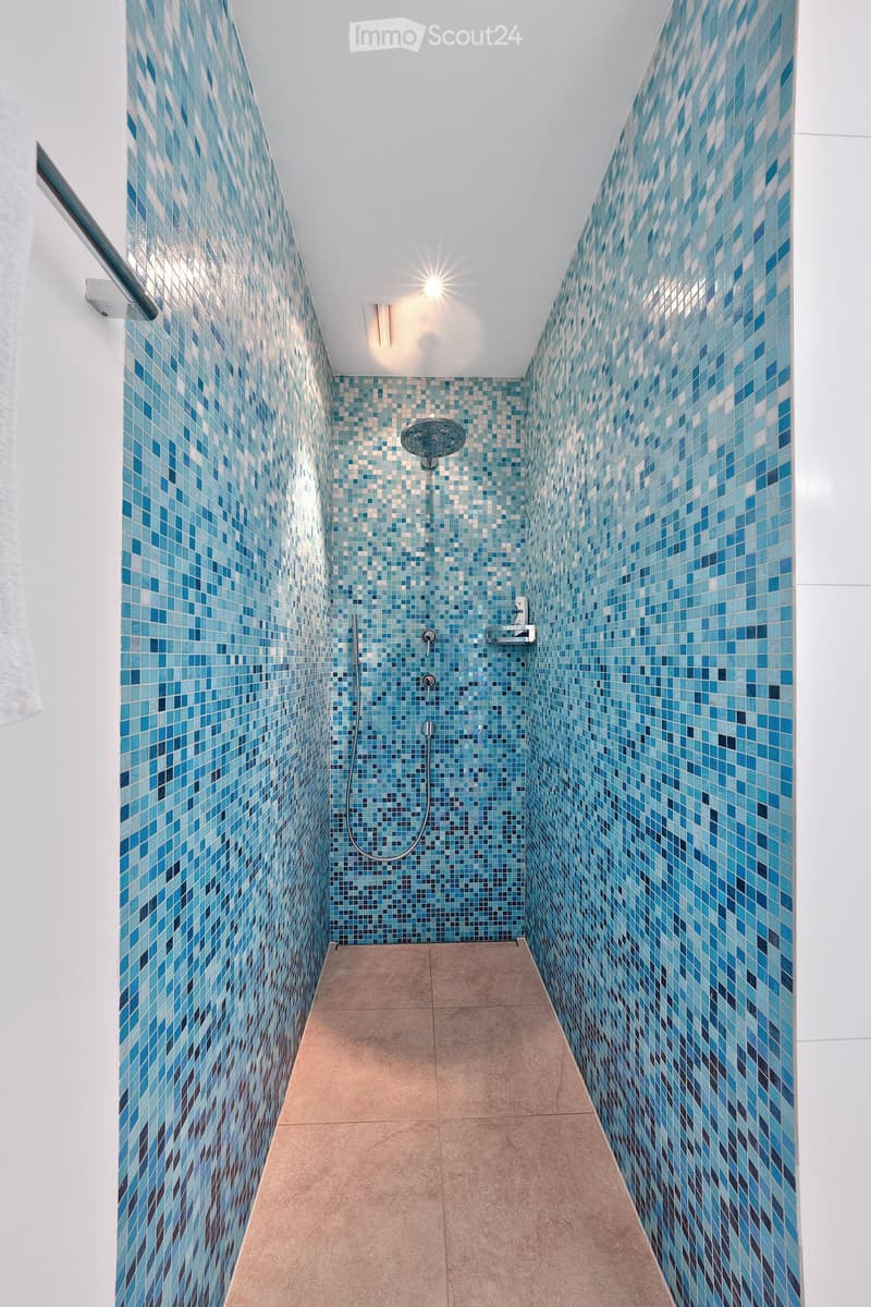 Modern shower area with blue mosaic tiles on walls, beige tiled floor, handheld showerhead, and built-in handrail.