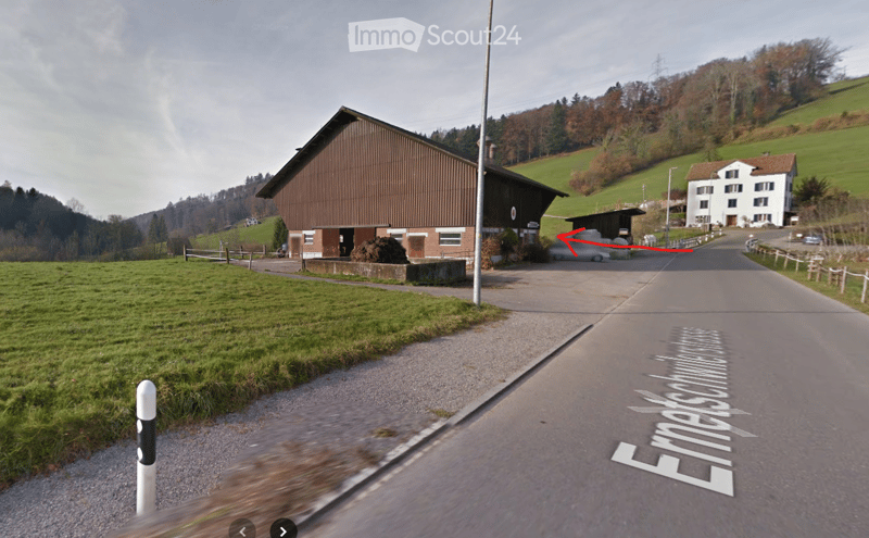A rural road scene with a large wooden barn on the left and a white house on the right, surrounded by green fields and hills.