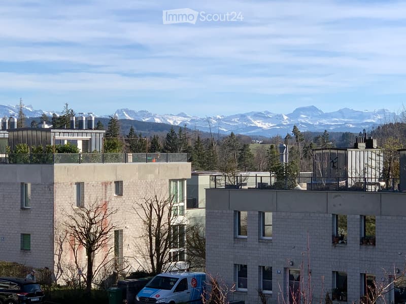 Modern multi-story residential building with balconies, surrounded by trees and mountains in the background.