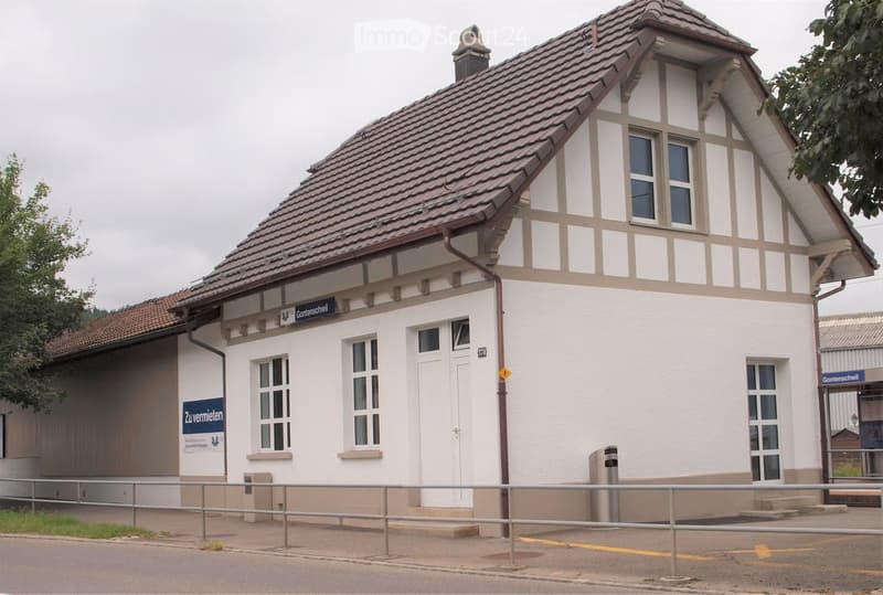 Two-story building with a tiled roof, white walls, and multiple windows. Features a sign reading "Gontenschwil" and a small entrance area with steps.