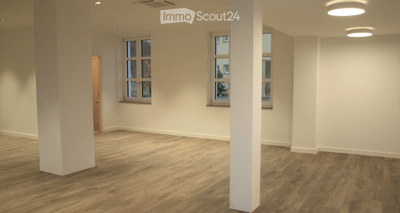 Empty room with light-colored walls, wooden flooring, and large windows with white frames. Minimalist design with a central column and recessed ceiling lighting.