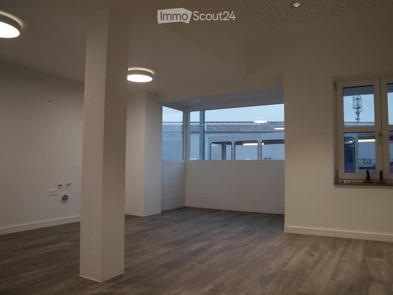 Empty room with wooden flooring, white walls, and large windows offering a view of an industrial area. The space is well-lit with recessed ceiling lights and features a central column.