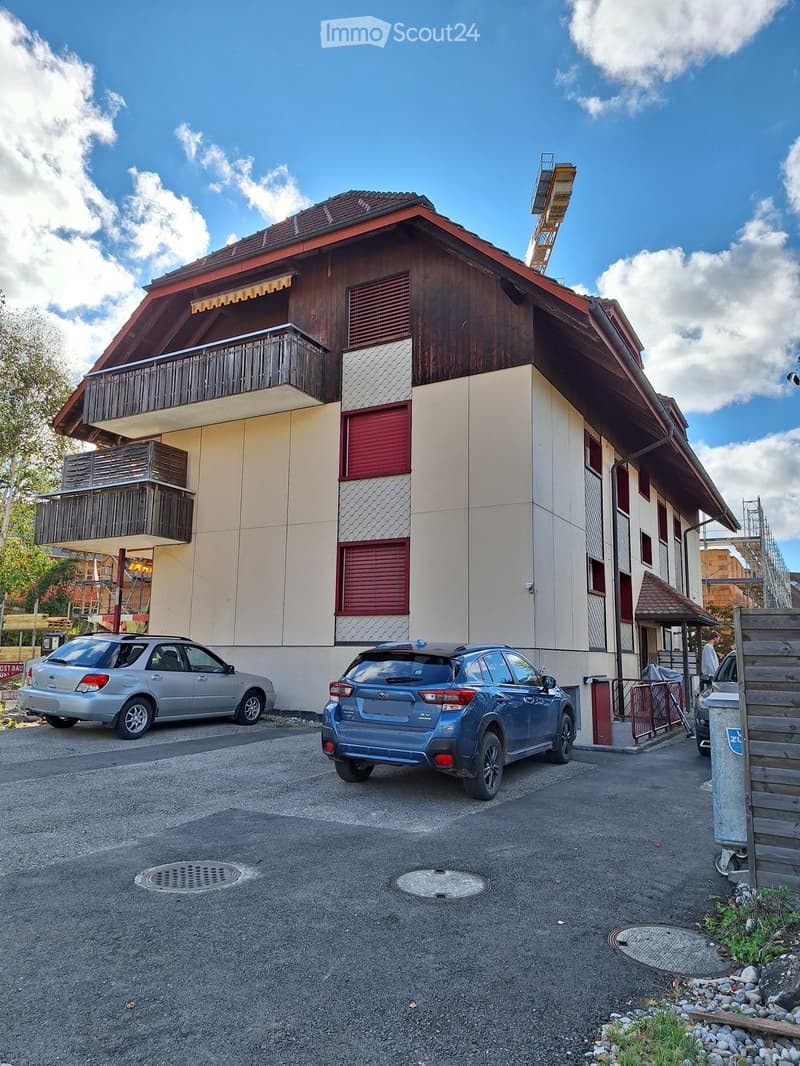 Three-story residential building, beige-brown exterior, red shutters, wooden balconies, cars parked