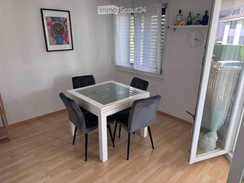 Dining room with a white dining table, four grey chairs, a clock on the wall, and a picture frame above the table. The room has wooden flooring and large windows with white shutters.
