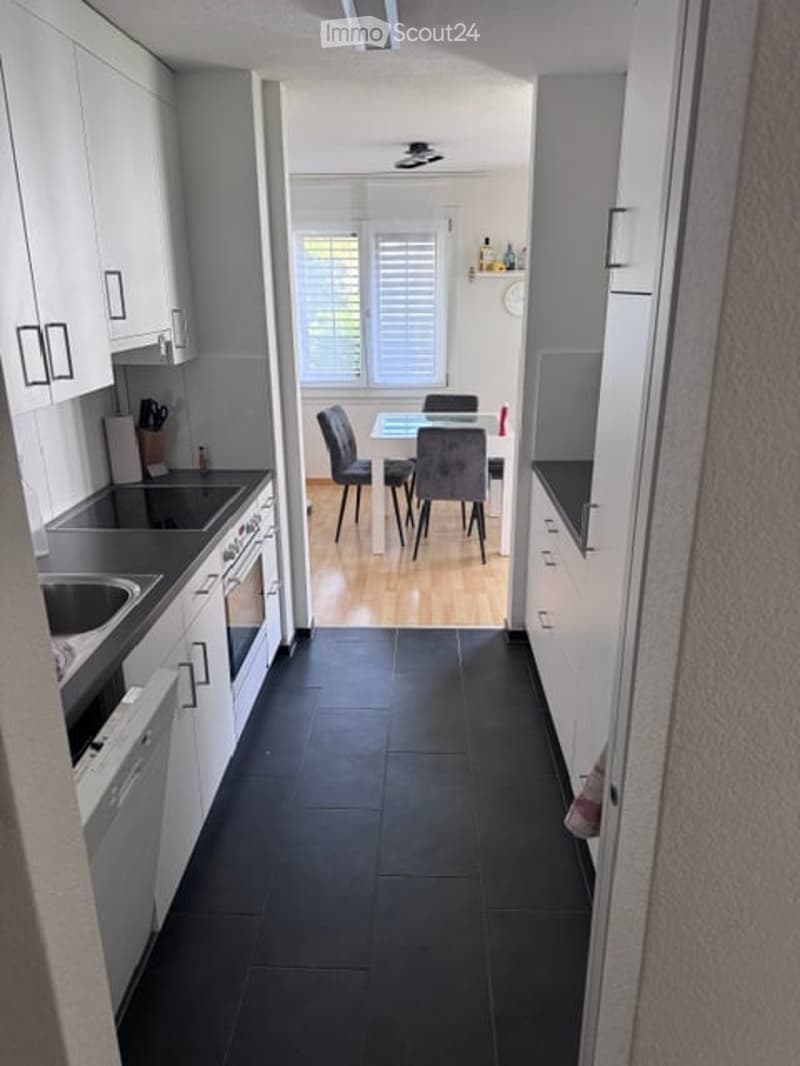 Modern kitchen with white cabinets, black countertops, and a dining area with a table and chairs. The floor is black tile, and the walls are light-colored.