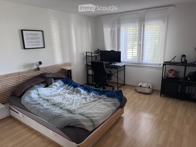 Cozy bedroom with a wooden bed, desk, chair, computer monitor, and shelves. The room features light wooden flooring and white walls.