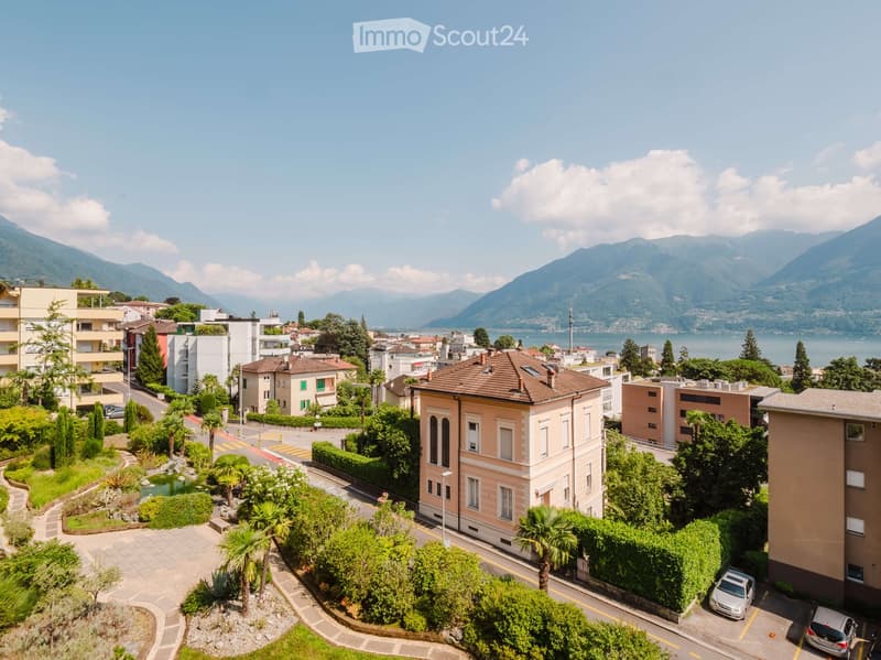 Aerial view of a residential area with a landscaped garden, palm trees, a small pond, and a road leading to a cluster of buildings with a scenic lake and mountains in the background.
