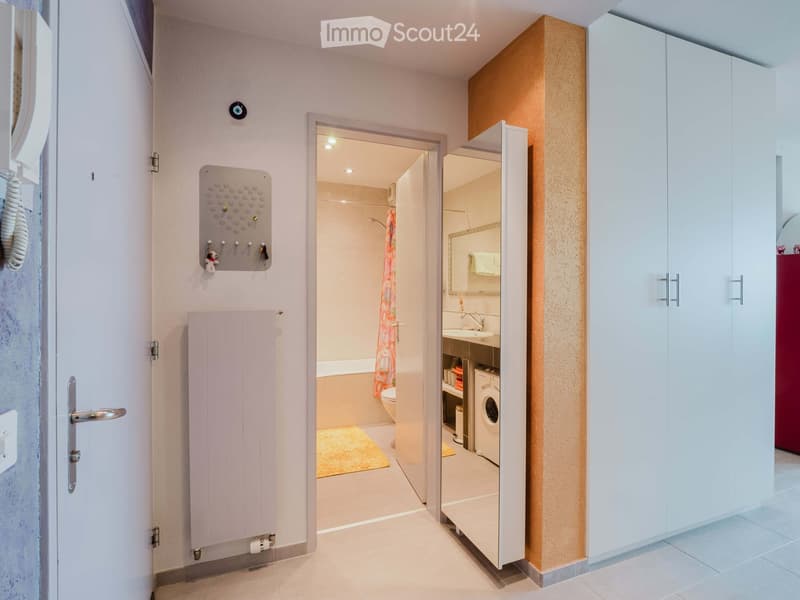 Hallway leading to a bathroom and a laundry area, featuring white cabinets, a radiator, and a coat rack with hooks.