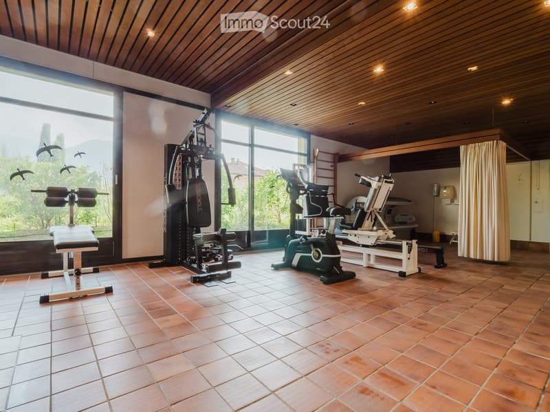Well-lit fitness room with wooden ceiling, tiled floor, and large windows overlooking a scenic outdoor area. Contains a bench, treadmill, elliptical machine, and multi-station gym machine.