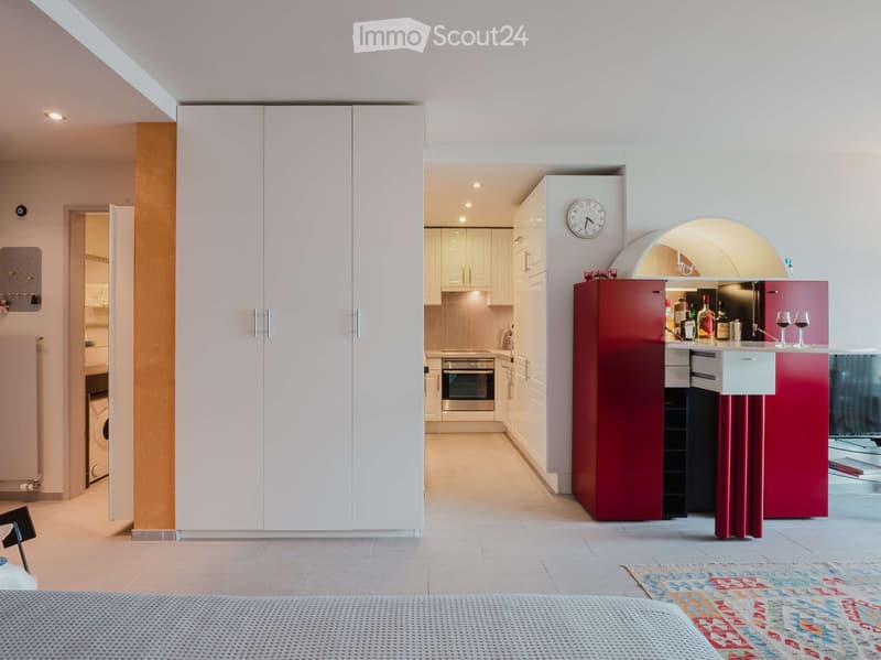 Modern, open-plan living space with a kitchen area featuring white cabinets, a red refrigerator, wine bottles, and wine glasses on the counter. A clock is mounted on the wall, and an oven and washing machine are visible in the background.