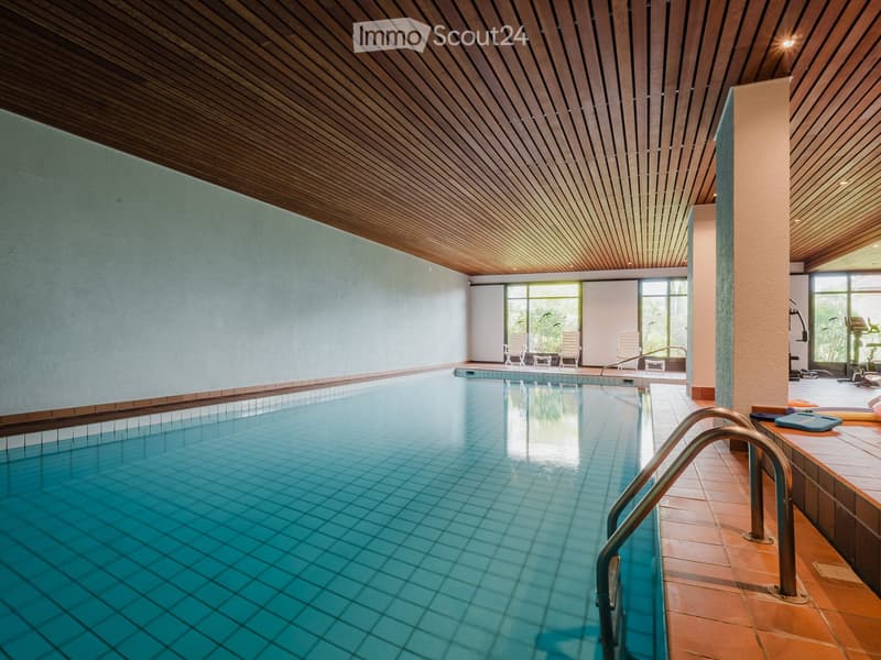 Indoor swimming pool area with wooden ceiling, tiled floor, and lounge chairs near large windows.