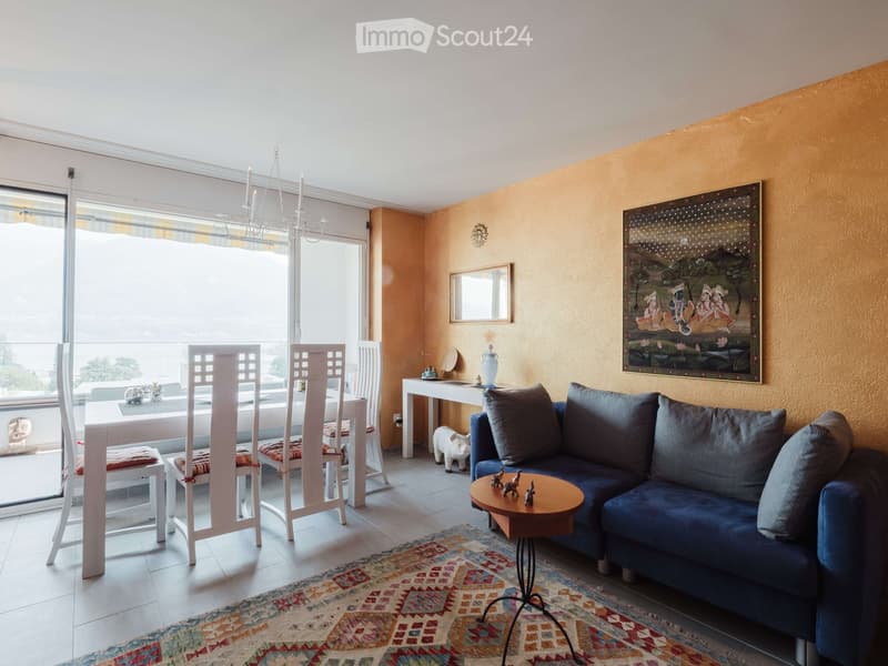 Bright living room with a blue sectional sofa, coffee table, dining area with white chairs, chandelier, and artwork on the walls.