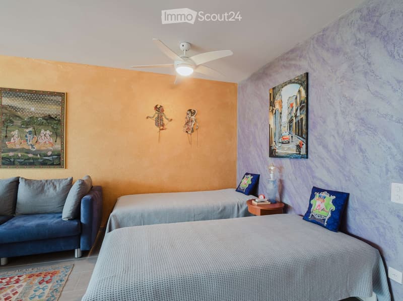 Cozy bedroom with two beds, a sofa, a small table with a lamp, and decorative pillows. The walls are painted in warm tones with artwork and a ceiling fan.