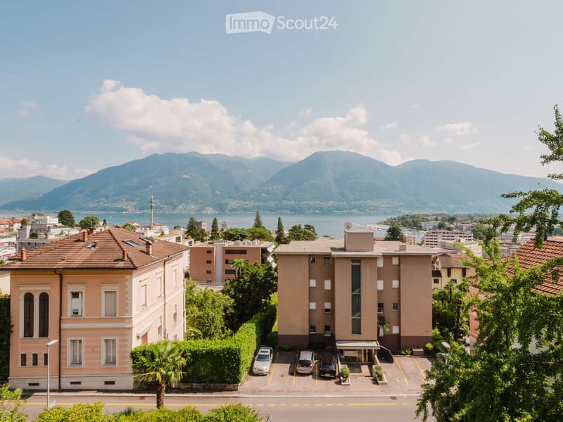 Multi-story residential building with a parking lot, surrounded by greenery and overlooking a lake with mountains in the background.