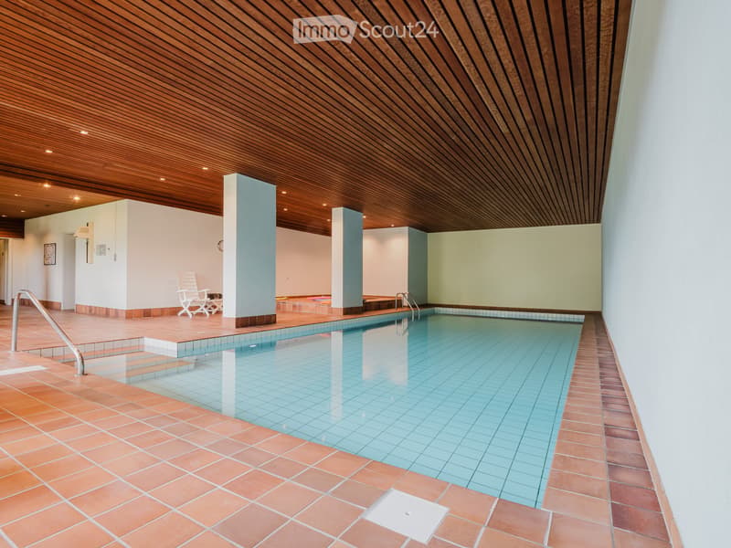 Indoor swimming pool area with tiled floors, wooden ceiling, lounge chairs, and a small seating area.