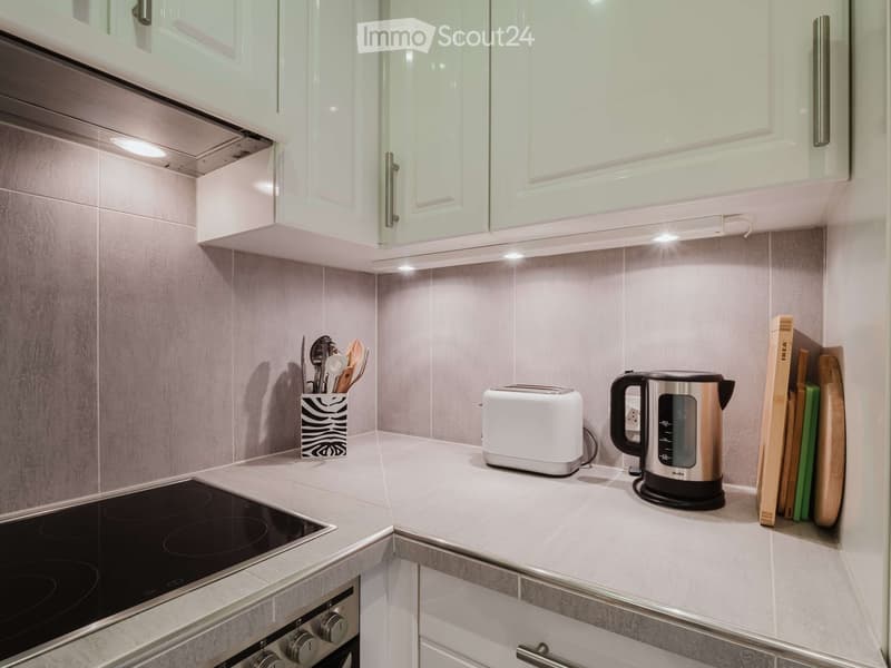 Modern kitchen with white cabinets, grey tiles, and stainless steel appliances including an electric kettle, toaster, and induction stove. Wooden cutting boards and a knife block are visible on the counter.