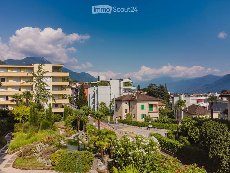 A scenic garden area with a small pond, surrounded by lush greenery and palm trees, with residential buildings and mountains in the background.