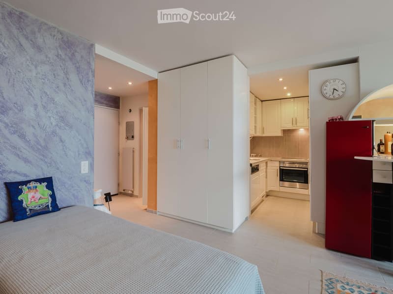 Modern bedroom with a neatly made bed, a large white wardrobe, and an open kitchen area featuring a red refrigerator, oven, and sink.