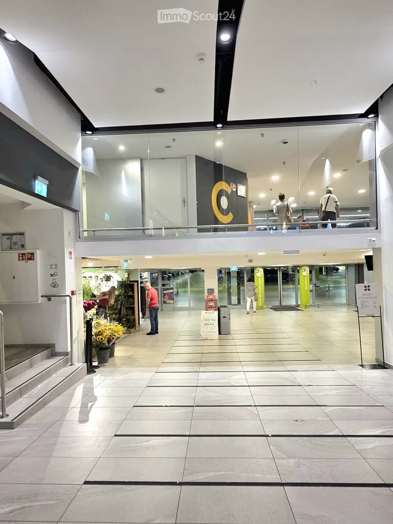 Modern interior hallway with tiled floor, glass walls, and a balcony level above. Features include potted plants, signage, and a staircase leading to an upper level.