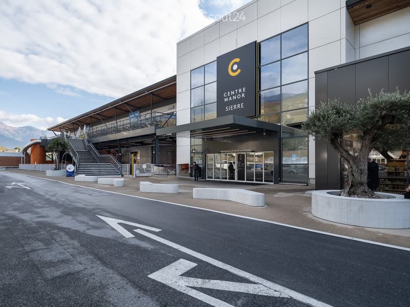 Modern building entrance with glass facade, sign reading "CENTRE MANOR SIERRE", steps leading up, and a tree in a planter.