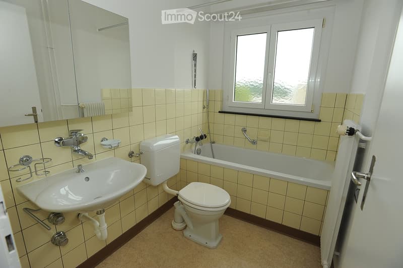 Bathroom with beige tiled walls, a white sink, toilet, bathtub, and a large mirror above the sink. The room has a window with frosted glass and a radiator below it.