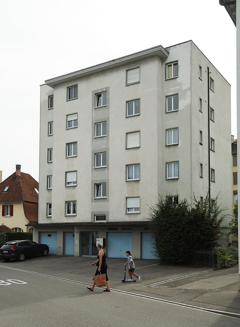 A multi-story residential building with light-colored walls and multiple windows, featuring a parking area in front with a car and two people walking.