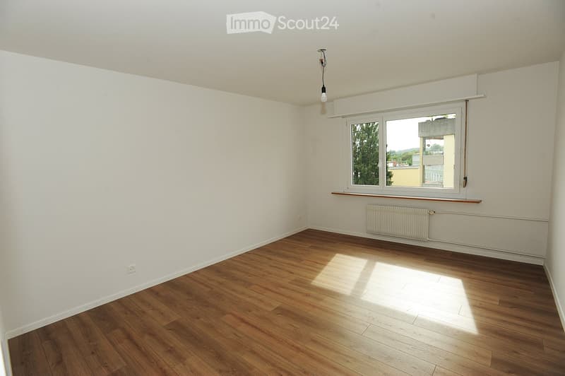 Empty room with wooden flooring, white walls, and a window with a radiator below it. Natural light streams in from the window, illuminating the space.