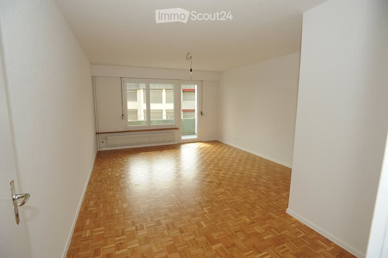 Empty room with wooden parquet flooring, white walls, large windows, and a radiator. No furniture or equipment present.