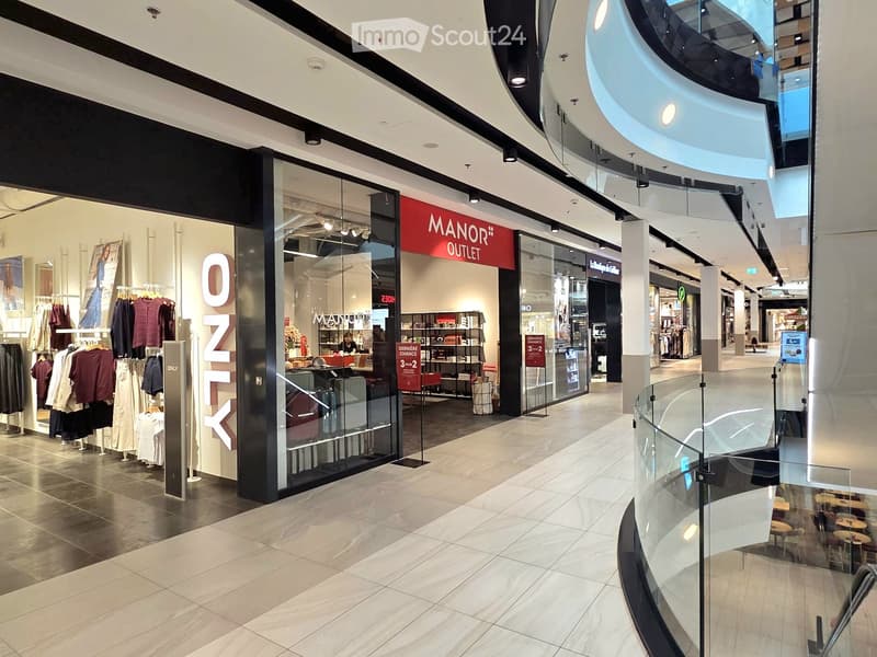 Shopping mall interior with glass storefronts, clothing racks, shelves, and a cashier counter visible inside a store.