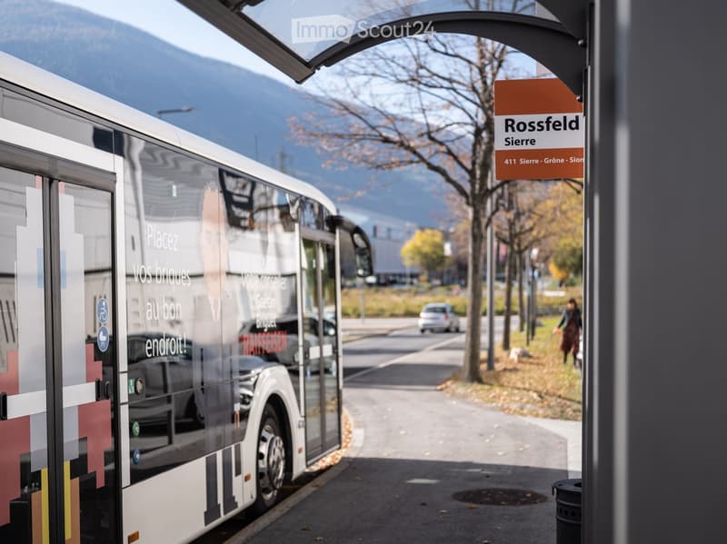 A modern bus is parked at a bus stop with a sign reading "Rossfeld Sierre." The background shows a scenic mountain view and a few trees.