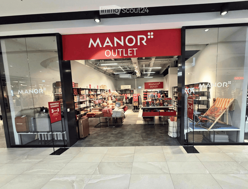 Storefront of Manor Outlet with glass windows displaying various items including luggage, shelves with folded towels, and a lounge chair.