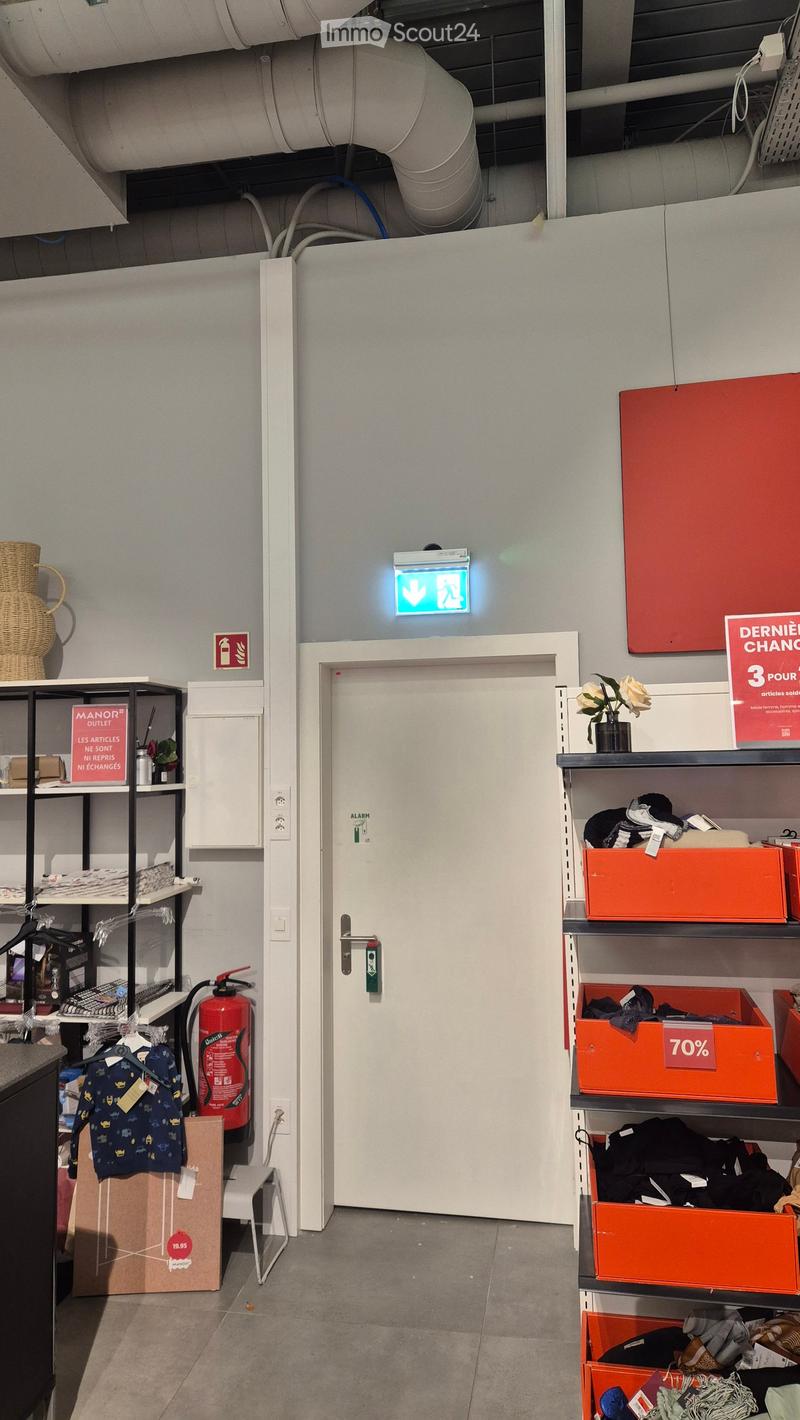 A well-lit hallway with a white door, shelves on both sides holding various items including clothing, boxes, and a fire extinguisher. A sign indicating an exit is visible above the door.