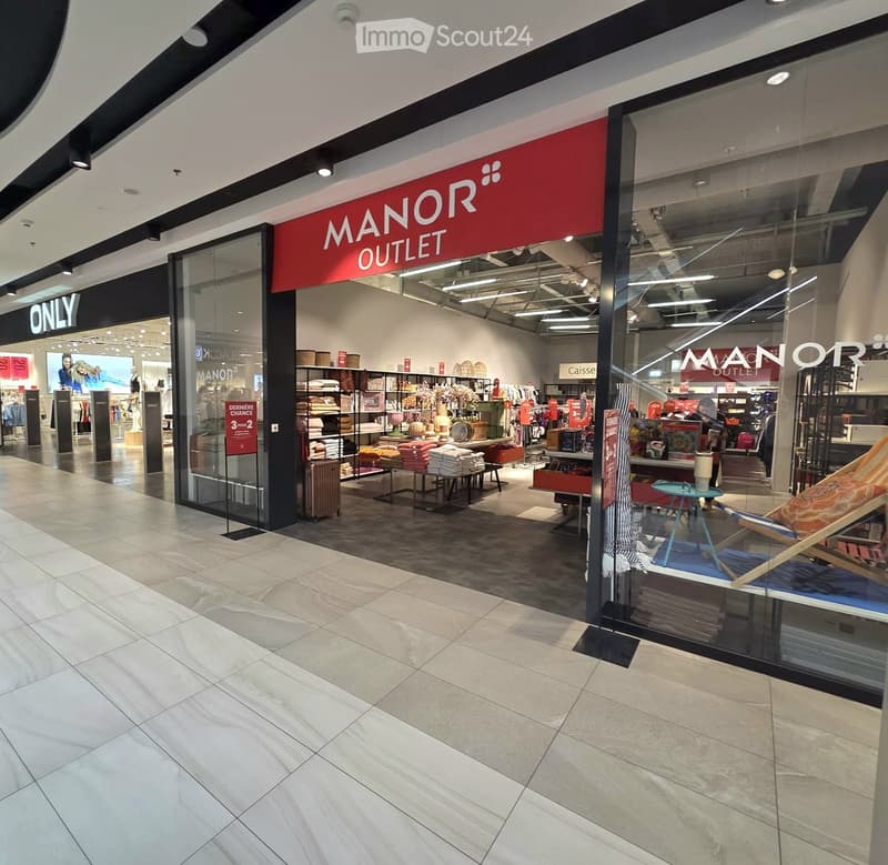 The image shows the entrance to a MANOR OUTLET store with glass windows displaying various items such as towels, a small table, a chair, and luggage. The store's interior is visible through the glass, showcasing shelves with folded towels and other mercha
