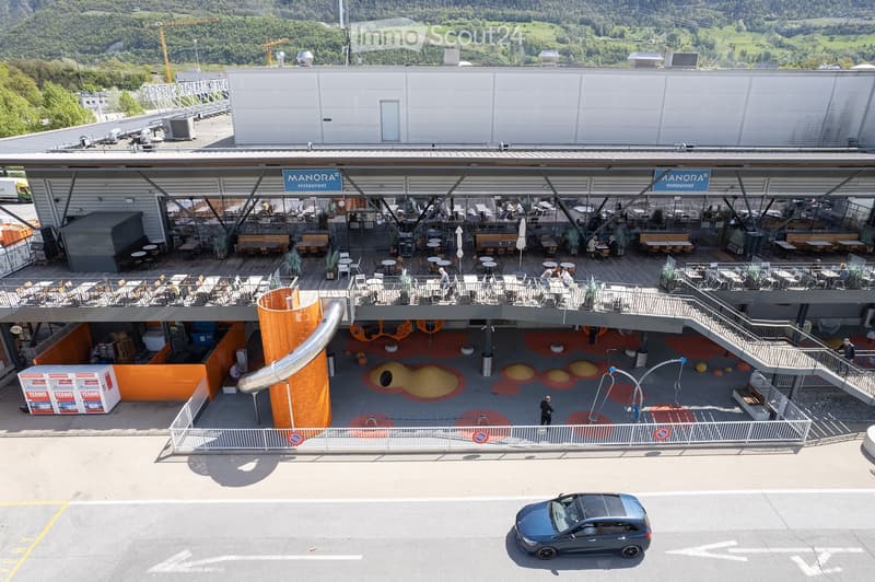 A modern building with a large outdoor seating area, featuring tables, chairs, and a restaurant sign. The area includes a colorful play area with a slide and other play structures.