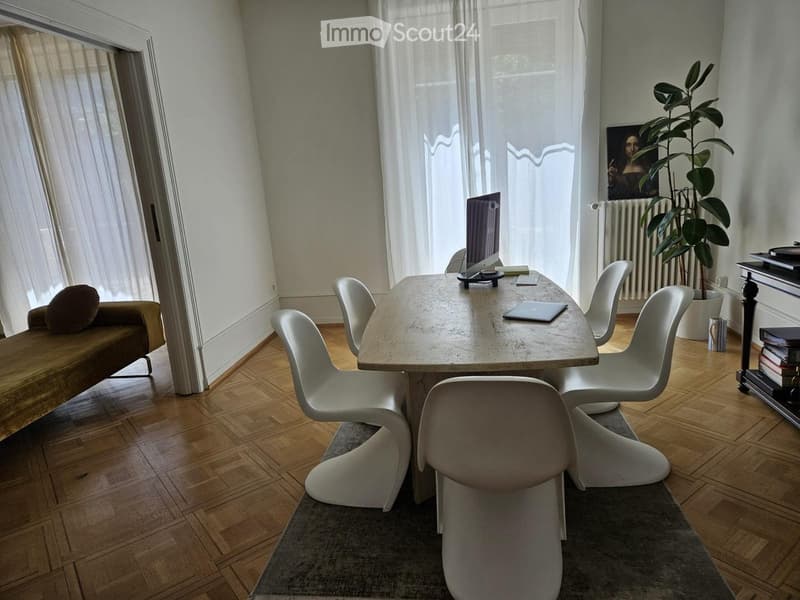 A modern, well-lit room with a wooden floor, a large window with sheer curtains, a white table surrounded by white chairs, a brown sofa, a laptop, a potted plant, and a picture frame on the wall.