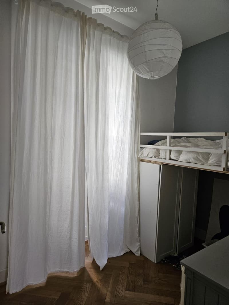 A small bedroom with a lofted twin-sized bed, white curtains, and a hanging pendant light. The room features wooden flooring and a built-in storage unit beneath the bed.