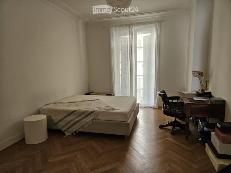 Bedroom with wooden floor, bed with striped blanket, desk with chair, lamp, and stool near the bed.