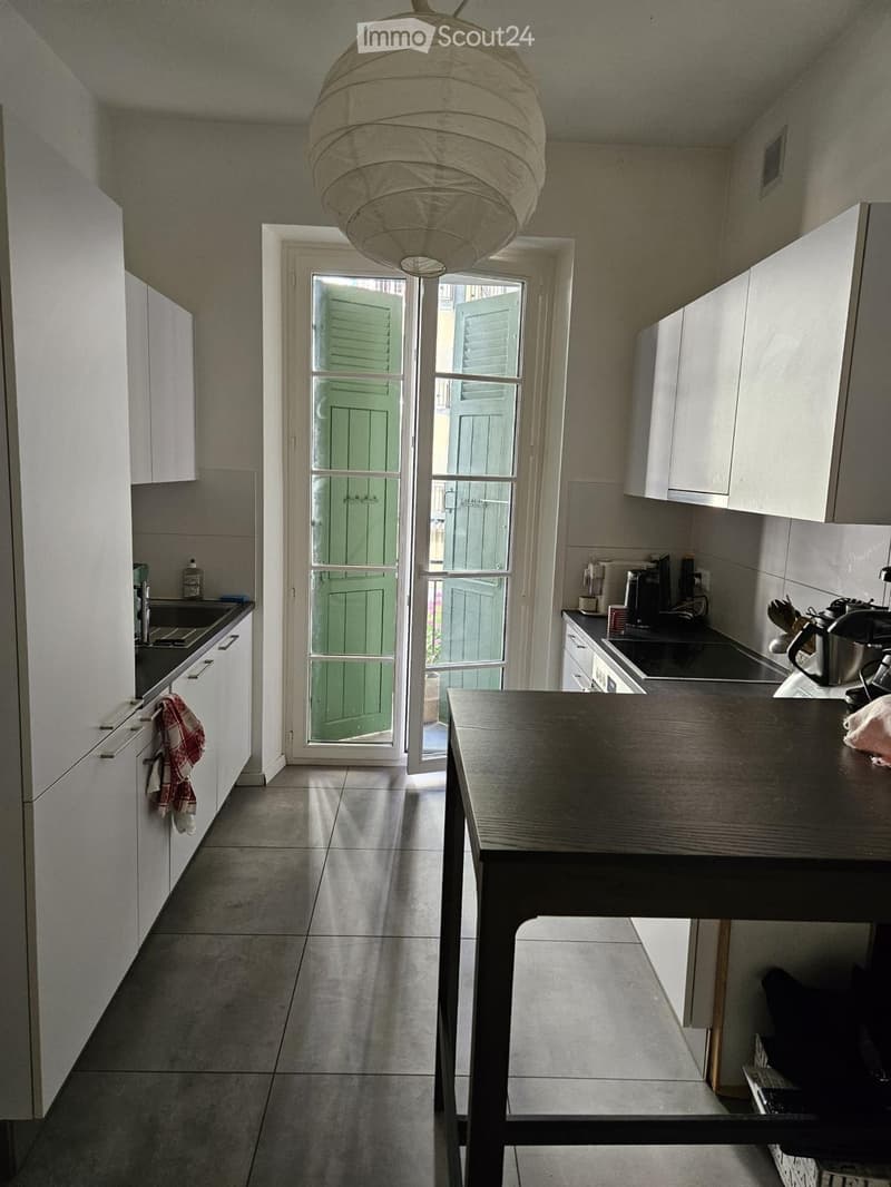 Modern kitchen with white cabinets, a central island, and a large window door leading outside. Visible items include a coffee maker, oven, sink, and a teapot on the counter.