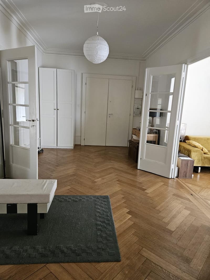 Spacious room with wooden herringbone flooring, a large sofa, a bed, a wardrobe, and a small table with a lamp.