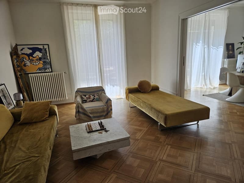 A well-lit living room with wooden flooring, large windows with white curtains, and a mix of modern and classic furniture including a sofa, chair, coffee table with a chessboard, and a desk area with a computer.