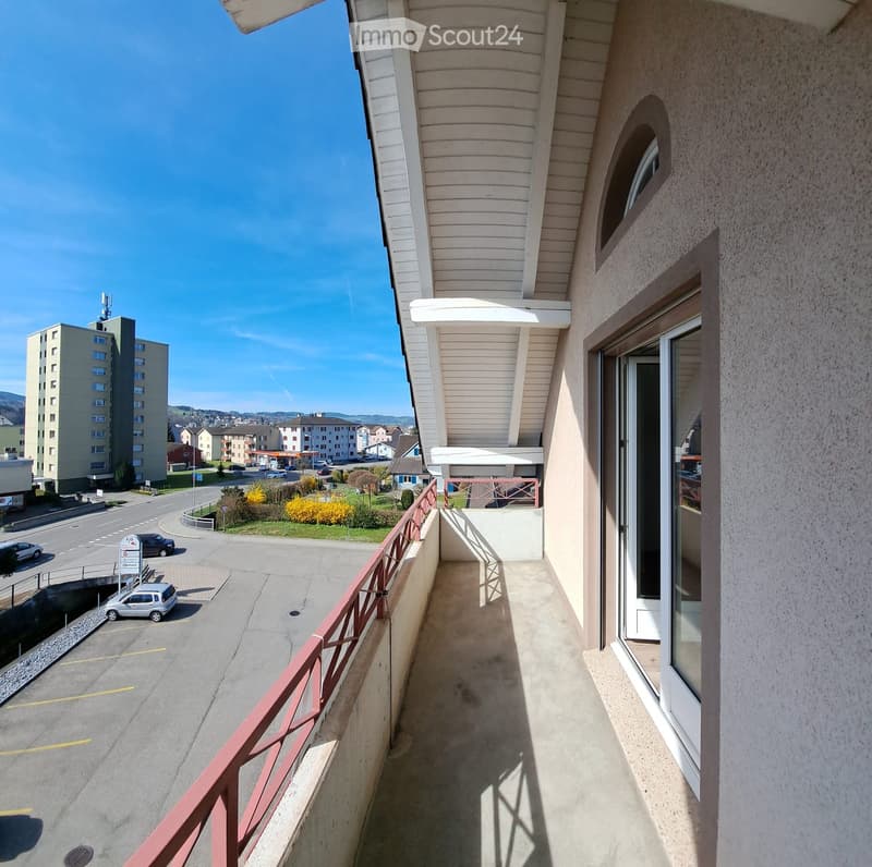 Balcony with a view of a street, parking area, and surrounding buildings. Features a red metal railing and a glass door leading inside.