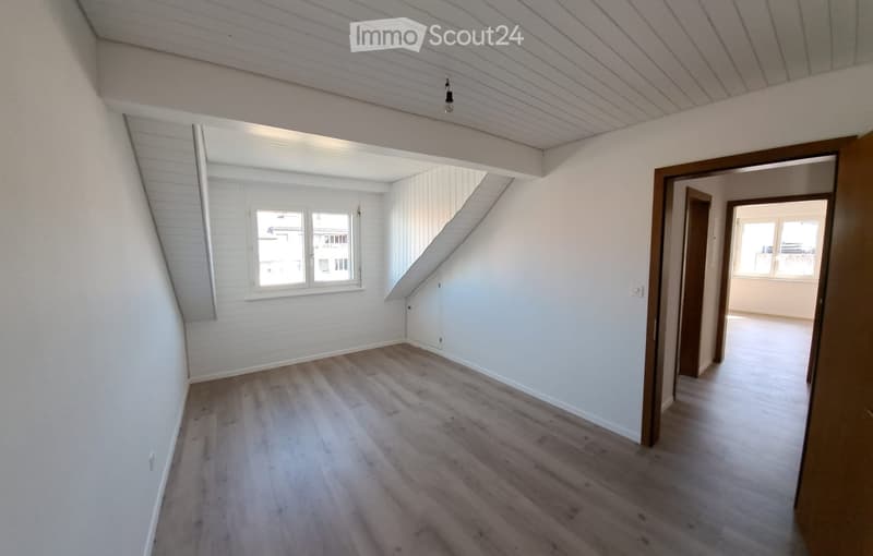 Empty room with wooden flooring, white walls, and a slanted ceiling. There are two windows and a doorway leading to another room.
