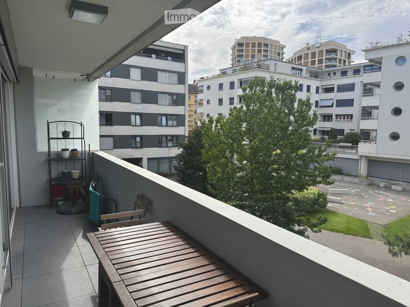 Balcony with wooden table and chairs, small shelf with pots, overlooking a residential area with modern buildings and greenery.